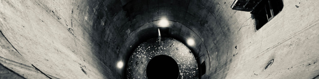 Tinted black and white photo, cropped into a horizontal banner format, looking down into a missile silo. It's a massive hollow cylinder made of concrete with some lights at the bottom and some open doorways in the walls.